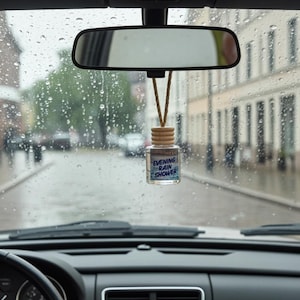 May include: A car air freshener with the text "Evening Rain Shower" hangs from the rearview mirror. The small glass bottle has a wooden cap and is suspended by a tan string. The car's interior is visible, with a rainy street scene in the background.
