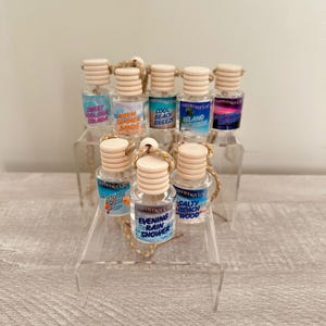 May include: A set of ten small glass bottles with cork stoppers and string hangers. Each bottle has a colorful label with a summer-themed scent name, such as "Sweet Poolside Colada", "Warm Summer Sands", "Cool Beach Breeze", "Island Vacation", "Orange Lush Pop", "Evening Rain Shower", and "Salty Beach Wood".