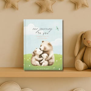 May include: A children's book titled "Our Journey to You" with a bedtime story of love and hope. The cover features a family of three bears hugging each other in a green grassy field with a blue sky and white clouds in the background.
