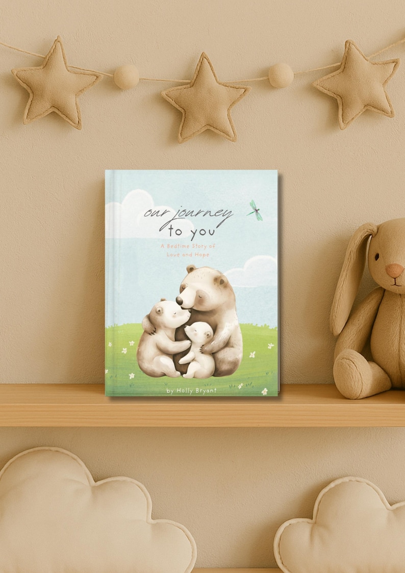 May include: A children's book titled "Our Journey to You" with a bedtime story of love and hope. The cover features a family of three bears hugging each other in a green grassy field with a blue sky and white clouds in the background.
