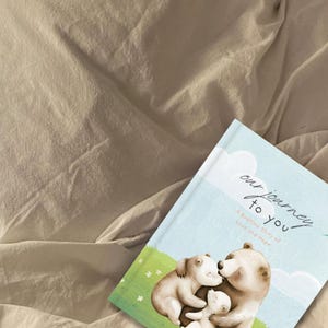 May include: A blue and white illustrated children's book titled "Our Journey to You: A Bedtime Story of Love and Hope" by Holly Bryant. The cover features a family of three bears hugging each other.