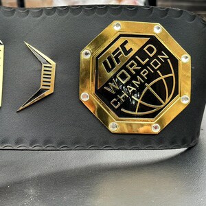 May include: A gold UFC World Champion belt with a black octagon-shaped logo and rhinestones. The belt is made of black leather with a textured surface.