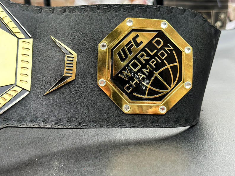 May include: A gold UFC World Champion belt with a black octagon-shaped logo and rhinestones. The belt is made of black leather with a textured surface.