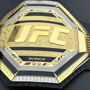 May include: A gold and silver UFC World Champion belt with black leather and diamond-like accents. The belt features the UFC logo and the words "World Champion" and "MCMXCIII".
