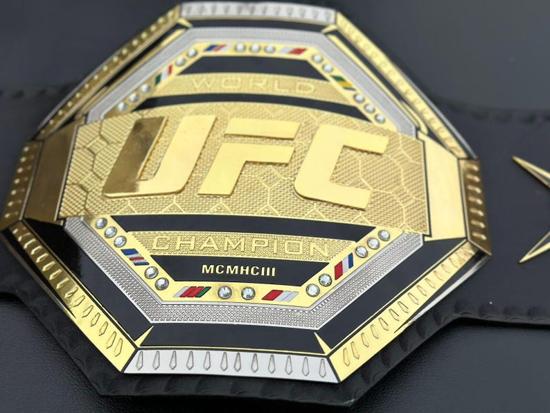 May include: A gold and silver UFC World Champion belt with black leather and diamond-like accents. The belt features the UFC logo and the words "World Champion" and "MCMXCIII".
