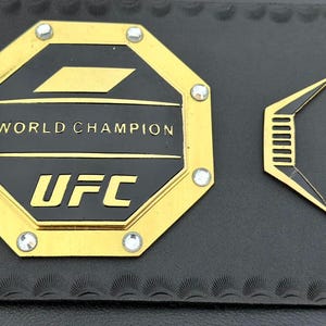 May include: A gold octagon-shaped belt buckle with the words "WORLD CHAMPION" and "UFC" in black lettering. The buckle is decorated with small rhinestones.