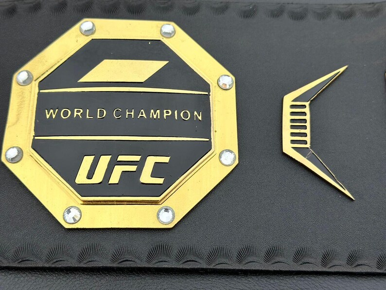 May include: A gold octagon-shaped belt buckle with the words "WORLD CHAMPION" and "UFC" in black lettering. The buckle is decorated with small rhinestones.