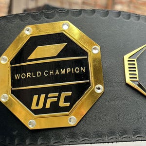 May include: A black leather belt with a gold octagonal buckle. The buckle features the words "World Champion" and "UFC" and is decorated with rhinestones.