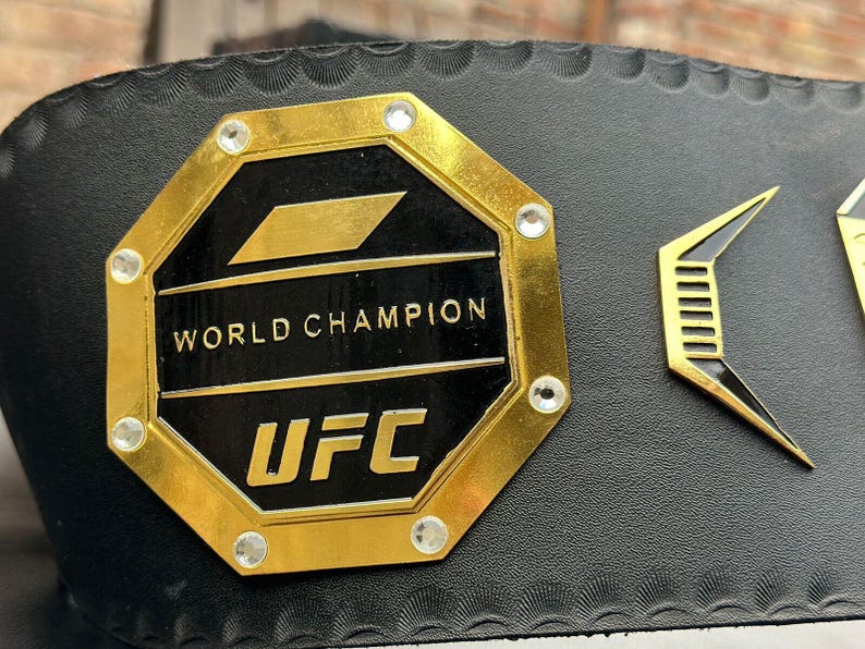 May include: A black leather belt with a gold octagonal buckle. The buckle features the words "World Champion" and "UFC" and is decorated with rhinestones.