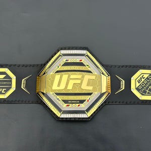 May include: A gold and black UFC World Champion belt with a large octagon-shaped buckle featuring the UFC logo and the words "World Champion".