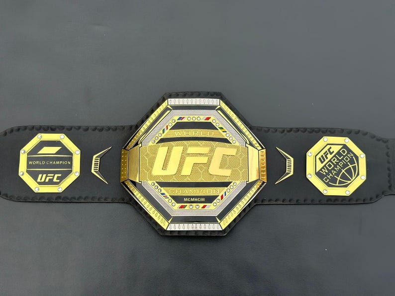 May include: A gold and black UFC World Champion belt with a large octagon-shaped buckle featuring the UFC logo and the words "World Champion".