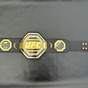 May include: A black leather UFC World Championship belt with gold accents. The centre plate features the UFC logo in gold lettering. The belt has a black strap with gold rivets.