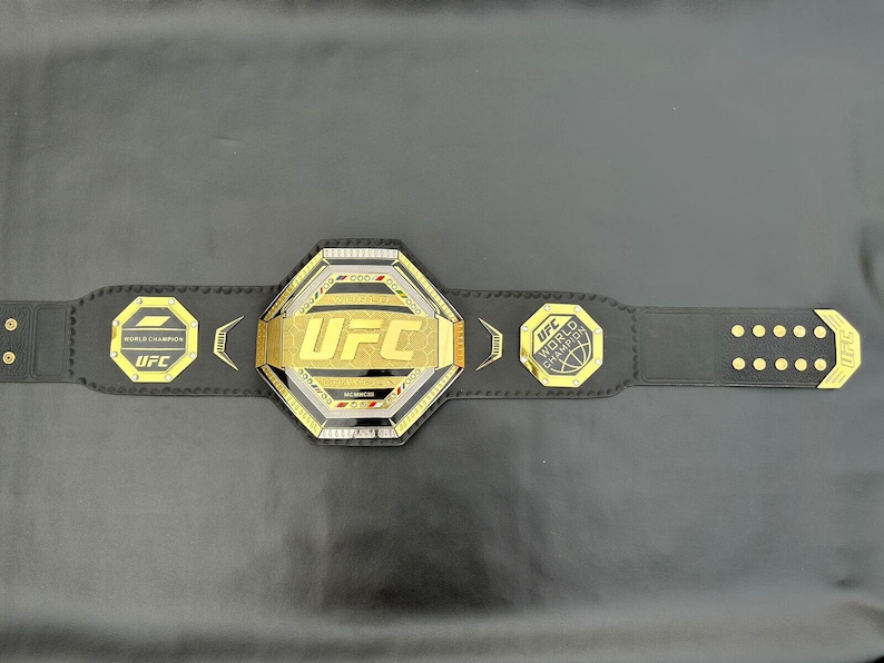 May include: A black leather UFC World Championship belt with gold accents. The centre plate features the UFC logo in gold lettering. The belt has a black strap with gold rivets.