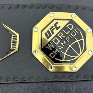 May include: A gold octagonal belt buckle with the text "UFC WORLD CHAMPION" and a black and gold globe design.