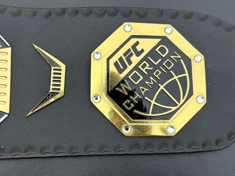 May include: A gold octagonal belt buckle with the text "UFC WORLD CHAMPION" and a black and gold globe design.