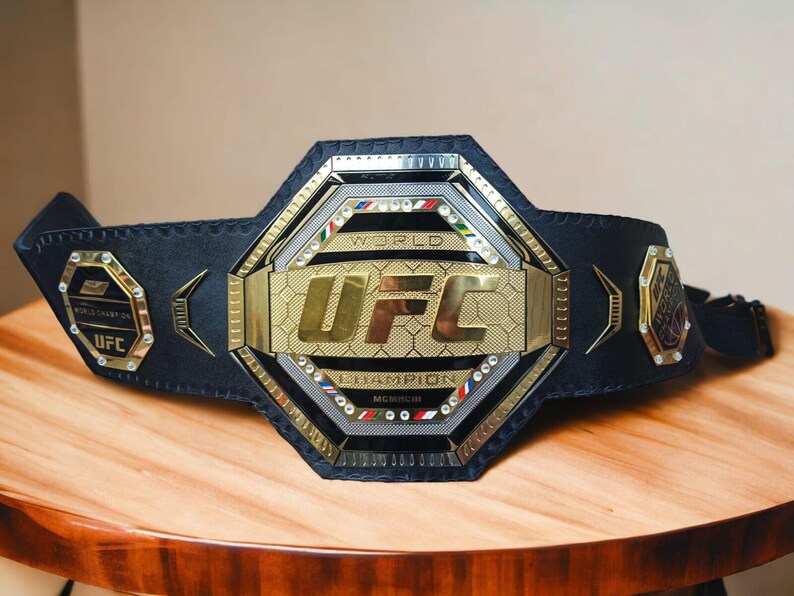 May include: A black leather UFC World Champion belt with a gold octagon-shaped buckle. The buckle features the UFC logo and the words "World Champion" in gold lettering.