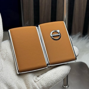 Puede incluir: Un estuche para llave de coche de cuero color marrón claro con un marco plateado. El logotipo de Volvo está centrado en la parte delantera. El estuche está abierto, revelando el interior. El estuche está sostenido por una mano enguantada.