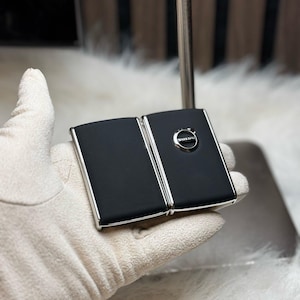 May include: A black, rectangular, folding key fob with a silver Volvo logo. The key fob is being held in a white glove.