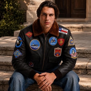 May include: A black leather bomber jacket with a brown faux fur collar, featuring numerous embroidered military-style patches including Texas Dallas, Seabees, and Tomcat, worn with blue denim jeans. The jacket has a front zipper and two pockets.