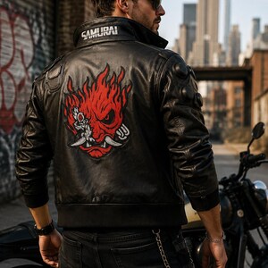 Könnte beinhalten: Eine schwarze Lederjacke mit einer großen rot-weißen Grafik einer stilisierten Oni-Maske auf dem Rücken und dem in Weiß auf den Kragen gestickten Wort SAMURAI. Die Jacke verfügt über gerippte Bündchen, einen gerippten Saum und kreisförmige Details.