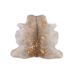 May include: A cowhide rug featuring a light beige and tan base colour with a metallic gold foil splatter pattern across the surface. The rug has an irregular, organic shape that mimics the natural silhouette of a hide with a central seam.