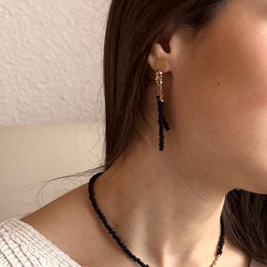 May include: A gold stud earring with a dangling black beaded branch-like charm and a matching necklace featuring a strand of small black beads accented with a section of gold beads, creating a coordinated jewelry set with a refined and simple aesthetic.