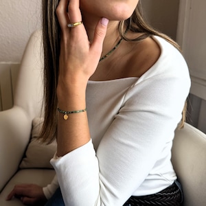 Op de afbeelding: Een gouden ring met een rechthoekige steen, een armband van groene kralen met een kleine ronde gouden bedel, en een ketting van groene kralen. De sieraden worden gedragen met een wit, geribd shirt met lange mouwen, een blauwe spijkerbroek en een donkerbruine gevlochten riem.