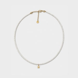 Peut inclure: Un collier de perles composé de petites perles cylindriques blanches uniformes. Le collier comprend une chaînette d'extension en métal doré avec une petite breloque circulaire et une étoile dorée suspendue au centre des perles.