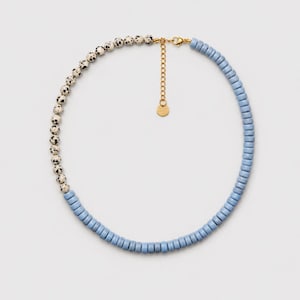 Può includere: Una collana dal design diviso, composta da un lato con perline rotonde color crema a pois neri e dall'altro con perline azzurre a forma di disco. Include una chiusura in metallo dorato e una catenella di estensione regolabile.