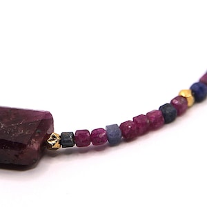 Könnte beinhalten: Eine Perlenkette mit einem großen, rechteckigen, facettierten dunkelroten Steinanhänger. Die Kette besteht aus kleinen, quadratisch geschliffenen Rubin- und Saphirperlen in Rot- und Blautönen, durchsetzt mit winzigen, polierten goldfarbenen Metallzwischenstücken.