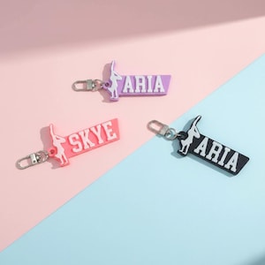 May include: Three keychains with a gymnast silhouette and name tags. One is pink with "SKYE" in white, another is purple with "ARIA" in white, and the last is black with "ARIA" in white. Each keychain has a silver clasp.