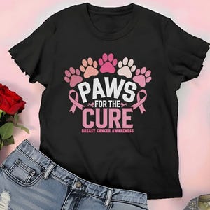 以下が含まれることがあります： 「PAWS FOR THE CURE」の文字とピンクと白の足跡がプリントされた黒いTシャツ。乳がん啓発を促進しています。Tシャツはデニムショーツ、赤いバラ、シルバーのハイヒールと組み合わせています。