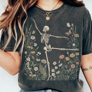 May include: A charcoal gray short-sleeve t-shirt featuring a graphic print of a human skeleton in a dancing pose surrounded by various wildflowers, ferns, and butterflies in muted tones of orange, yellow, and white across the front chest area.