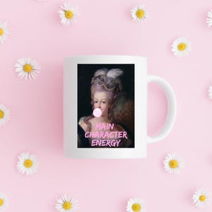 May include: White ceramic mug featuring a portrait of a woman with a bubblegum bubble, with the text "MAIN CHARACTER ENERGY" in pink. The mug is surrounded by daisies on a pink background.