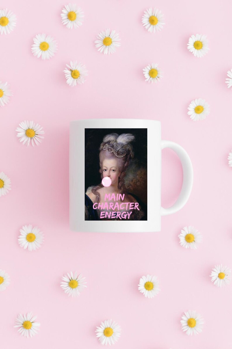 May include: White ceramic mug featuring a portrait of a woman with a bubblegum bubble, with the text "MAIN CHARACTER ENERGY" in pink. The mug is surrounded by daisies on a pink background.