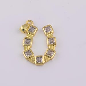 May include: A gold-colored horseshoe charm with square, clear gemstones set in each section. The charm has a small loop at the top for hanging and a small heart-shaped accent. The charm is likely for jewelry making.