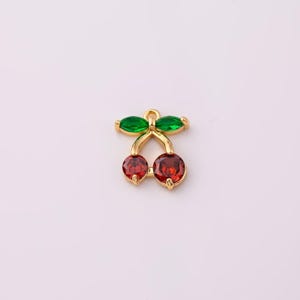 Op de afbeelding: Een goudkleurige kersenbedel met rode edelsteenkersen en groene edelsteenbladeren. De bedel heeft een lus aan de bovenkant om aan sieraden te bevestigen. De kersenbedel is een schattig accessoire.