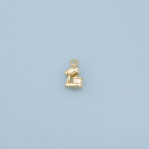 May include: A small, polished gold-tone charm shaped like a stand mixer. The charm features a rounded top section, a central arm, and a cylindrical base representing the mixing bowl, with a small circular jump ring attached to the top for hanging.