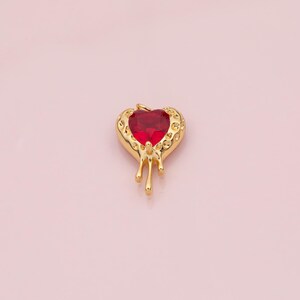 May include: A gold heart pendant with a red gemstone centre. The pendant has a textured border and a dripping design. The background is a light pink colour. The pendant is likely a piece of jewellery.