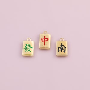 May include: Three gold-colored rectangular charms with Chinese characters in red, green, and black. Each charm has a small loop at the top for attaching to a chain or bracelet. The background is a light purple color.