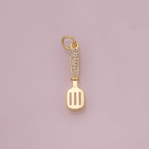 May include: A gold-coloured pendant shaped like a spatula. The handle is covered in small, clear crystals. The spatula head has three vertical slots. The pendant has a small loop at the top for a chain.