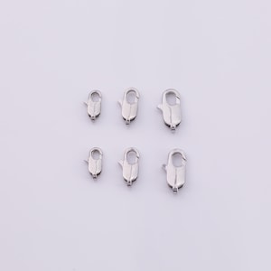 Puede incluir: Seis cierres de pinza de langosta plateados de diferentes tamaños están dispuestos sobre un fondo blanco. Estos cierres se utilizan en la fabricación de joyas para collares y pulseras. Los cierres son de metal brillante.