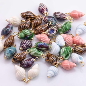 May include: A collection of ceramic seashell charms in various colors, including white, pink, green, blue, brown, and purple. Each charm has a gold-colored loop for attaching to jewelry or other crafts. The seashells are detailed with textured surfaces.