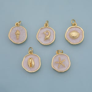 Op de afbeelding: Vijf goudkleurige hangers met witte emaille achtergronden en verschillende zeethema-ontwerpen. De hangers hebben een zeeschelp, een dolfijn, een sint-jakobsschelp, een kaurischelp en een zeester. Elke hanger heeft een gouden lus.