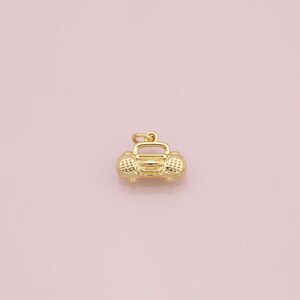 Könnte beinhalten: Ein goldfarbener Anhänger in Form einer Retro-Boombox. Der Anhänger hat detaillierte Lautsprecher- und Knopfanordnungen mit einer Schlaufe oben zum Befestigen an einer Halskette oder einem Armband. Der Anhänger befindet sich vor einem weichen rosa Hintergrund.