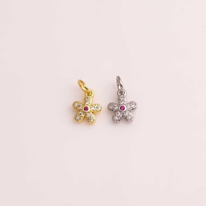 May include: Two flower-shaped charms. One is gold-colored with clear and red accents, the other is silver-colored with clear and red accents. Each charm has a small loop at the top for attaching to a necklace or bracelet. The charms are small and delicate.