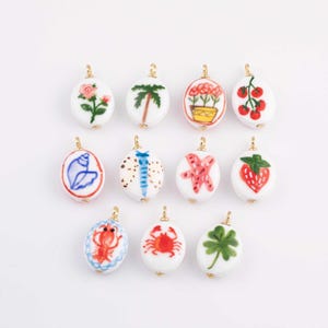 May include: A collection of white oval-shaped ceramic pendants with gold-colored loops. Each pendant features a hand-painted design, including a rose, palm tree, flowers, tomatoes, seashell, butterfly, starfish, strawberry, lobster, crab, and a four-leaf clover.