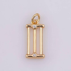Pode incluir: Pingente dourado com design retangular. O pingente apresenta duas barras verticais com borda serrilhada e uma barra central. Pequenas pedras preciosas transparentes são colocadas na parte superior e inferior. Uma pequena argola na parte superior.