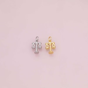 May include: Two small pendants shaped like scales of justice. One is silver-colored, and the other is gold-colored. The pendants are on a light pink background. The scales have a loop at the top for attaching to a chain or bracelet.