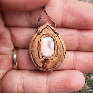 Puede incluir: Un colgante de madera con un diseño tallado y una incrustación de piedra blanca. El colgante está unido a un cordón marrón.